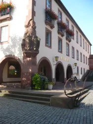 Ein historisches Gebäude mit einer Statue und blühenden Fensterkästen. Der Fußweg ist aus Pflastersteinen und bietet einen einladenden Eingang.