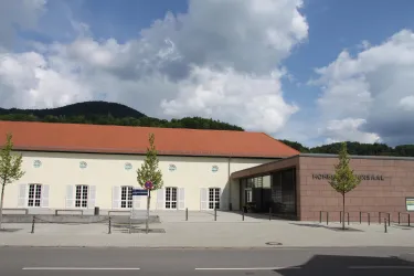 Ein modernes Gebäude mit einem roten Dach und großen Fenstern. Im Hintergrund sieht man hügelige Landschaften und einen klaren Himmel.