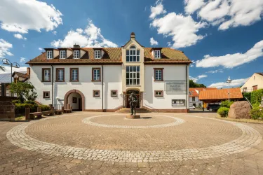 Ein historisches Gebäude mit einem Geschmack von Tradition und Modernität, das von einer runden Pflasterfläche umgeben ist. Strahlend blauer Himmel und ein paar Wolken komplettieren die Szenerie.