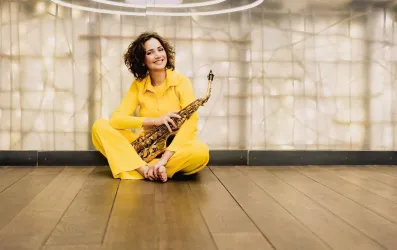 Eine lächelnde Frau sitzt auf dem Boden und hält ein Saxophon in der Hand. Sie trägt einen gelben Anzug und hat lockige Haare.