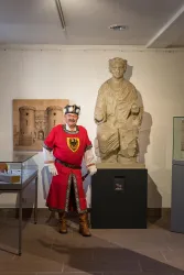 Ein Mann in einer roten Ritterrüstung steht neben einer Statue in einem Museum. Im Hintergrund sind weitere Ausstellungsstücke zu sehen.