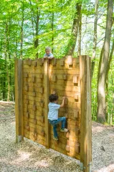 Ein Kletterspielplatz im Wald mit zwei Kindern. Eines der Kinder klettert die Wand hinauf, während das andere darüber sitzt.