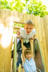 Zwei Kinder spielen auf einer Rutsche in einem Spielplatz. Die Sonne scheint durch die Bäume und sorgt für eine fröhliche Atmosphäre.