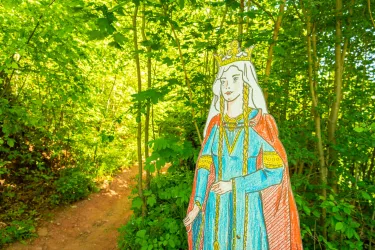 Eine bunte Pappfigur einer Königin steht im Wald. Der Weg im Hintergrund ist von grünen Bäumen umgeben.