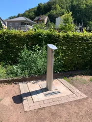 Eine moderne Wasserstation aus Edelstahl steht in einer grünen Umgebung. Im Hintergrund sind Wohnhäuser und Bäume sichtbar.