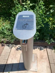 Ein Trinkwasserspender aus Edelstahl steht auf einem gepflasterten Weg. Im Hintergrund sind grüne Pflanzen zu sehen.