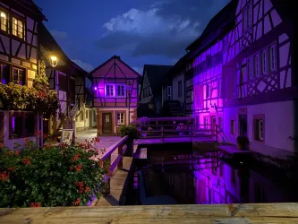 Eine malerische Altstadt mit Fachwerkhäusern und einem kleinen Wasserlauf. Die Szene wird von lila Beleuchtung und einem stimmungsvollen Himmel bei Nacht geprägt.