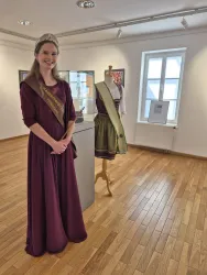 Eine Frau in einem burgunderfarbenen Kleid steht lächelnd in einem Ausstellungsraum. Im Hintergrund ist ein Kleid auf einer Schneiderpuppe zu sehen.