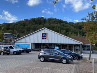 Ein Aldi-Supermarkt mit einem modernen Gebäude. Es sind mehrere Autos auf dem Parkplatz zu sehen, und im Hintergrund befinden sich Bäume.