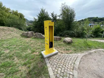 Eine gelbe Telefonzelle steht in einer grünen Umgebung. Im Hintergrund sind Bäume und ein bewölkter Himmel zu sehen.