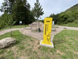 Eine Radservice-Station mit einem gelben Schild steht neben einer Wiese. Im Hintergrund sind Bäume und ein Weg zu sehen.