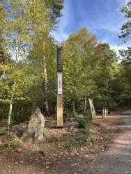 Ein Waldweg mit Bäumen und einer Informationssäule. Auf der linken Seite steht ein großer Felsen mit der Aufschrift „Fritz“.