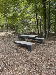 Eine moderne Sitzbank aus Holz und Metall steht im Wald. Umgeben von Bäumen und Laub wirkt die Bank einladend und gemütlich.