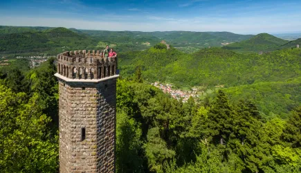Ein steinerner Turm steht auf einem Hügel, umgeben von grünen Wäldern und sanften Bergen. Im Hintergrund ist eine malerische Landschaft mit einem kleinen Dorf zu sehen.