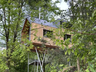 Ein modernes Baumhaus aus Holz, umgeben von grünen Bäumen. Es steht auf Stelzen und hat große Fenster.