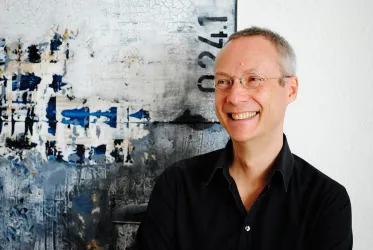 Ein lächelnder Mann mit einer Brille steht vor einer abstrakten, künstlerischen Wandgestaltung. Die Farben sind hauptsächlich Blau und Beige.