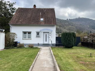 Ein kleines Haus mit einem gepflegten Vorgarten. Im Hintergrund sind sanfte Hügel und ein bewölkter Himmel zu sehen.