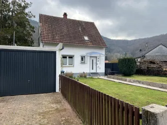 Ein kleines, weißes Haus mit einem Satteldach und einer Garage. Der Garten ist gepflegt und es gibt einen Blick auf die umliegenden Hügel.