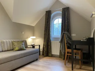Ein gemütliches Zimmer mit schrägem Dach. Es gibt ein Sofa, einen Schreibtisch und ein Fenster mit Vorhängen.