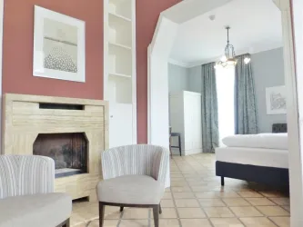 Ein helles Zimmer mit zwei beigen Stühlen und einem Kamin. Im Hintergrund sind ein Bett und große Fenster mit Vorhängen zu sehen.