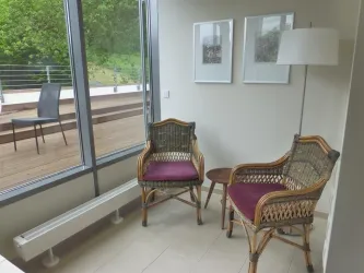 Ein gemütlicher Sitzbereich mit zwei Rattanstühlen und einem kleinen Tisch. Große Fenster bieten einen Blick auf eine Holzterrasse und die grüne Umgebung.