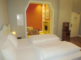 Ein gemütliches Schlafzimmer mit zwei Betten und großen Kopfkissen. Im Hintergrund ist ein weiterer Bereich mit einem Sessel und einem Kamin sichtbar.