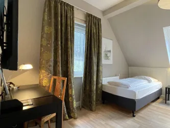 Ein gemütliches Schlafzimmer mit einem Bett und einem Schreibtisch. Große Vorhänge lassen viel Licht herein und schaffen eine einladende Atmosphäre.