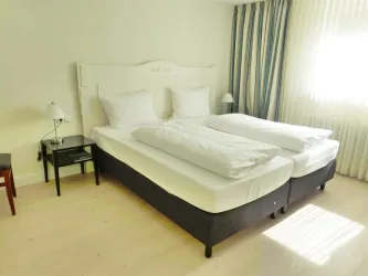 Ein helles und modernes Zimmer mit zwei Betten und großen Fenstern. Neben dem Bett befindet sich ein Nachttisch und ein Stuhl.