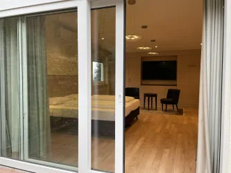 Ein modernes Zimmer mit einem Fenster und einer Tür aus Glas. Im Inneren sind ein Bett, ein Tisch und zwei Stühle sichtbar.