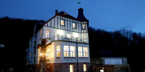 Ein charmantes Haus mit einem Turm, beleuchtet in der Dämmerung. Im Hintergrund sind Bäume und ein klarer Himmel zu sehen.