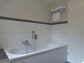 Ein modernes Badezimmer mit einer weißen Badewanne und grauen Fliesen. Saubere Handtücher hängen an einem Regal über der Badewanne.