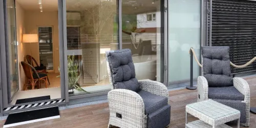 Ein gemütlicher Außenbereich mit zwei bequemen Stühlen und einem kleinen Tisch. Der Blick führt auf einen hellen Innenraum mit Pflanzen und einer warmen Beleuchtung.