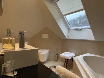 Ein modernes Badezimmer mit schrägem Fenster und einer Badewanne. Auf dem Waschbecken stehen Seifen und Handtücher.