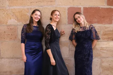 Drei Frauen in eleganten, dunkelblauen Kleidern posieren vor einer Steinmauer. Sie lächeln und haben unterschiedliche Frisuren.