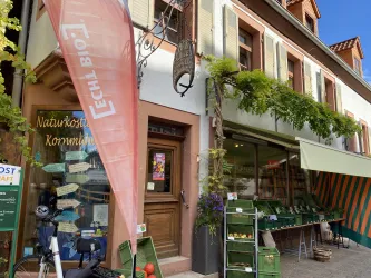 Ein wunderschöner Markt mit frischem Obst und Gemüse vor einem gemütlichen Laden. Die Fassade des Gebäudes ist hell und einladend, mit einem klaren blauen Himmel im Hintergrund.