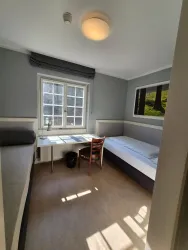 Ein einfaches Zimmer mit zwei Betten und einem Arbeitsplatz. Der Raum hat ein Fenster, das Tageslicht hereinlässt.