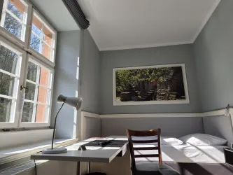 Ein gemütliches Zimmer mit einem Einzelbett und einem Schreibtisch. Große Fenster lassen viel Licht herein und zeigen eine grüne Umgebung.