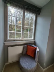 Eine gemütliche Sitzecke an einem Fenster mit Blick auf einen Wald. Der Sessel hat ein rotes Kissen und der Raum ist hell und einladend.