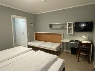 Ein einfaches Zimmer mit zwei Betten und einem Schlafsofa. Es gibt einen Schreibtisch und einen Fernseher an der Wand.