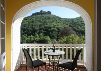 Ein schöner Balkon mit einem kleinen Tisch und zwei Stühlen. Im Hintergrund ist eine grüne Hügellandschaft mit einer Burg zu sehen.