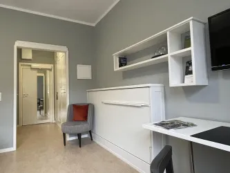 Ein modernes Zimmer mit grauen Wänden und einem praktischen Wandbett. Der Raum verfügt über einen Schreibtisch und eine kleine Sitzecke.