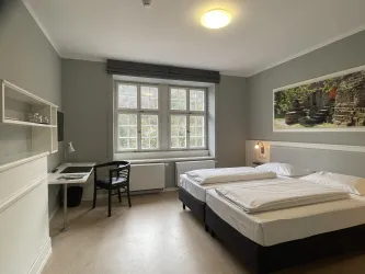 Ein modernes Zimmer mit zwei Betten und einem Schreibtisch. Große Fenster bieten viel Tageslicht und einen Blick auf die Umgebung.