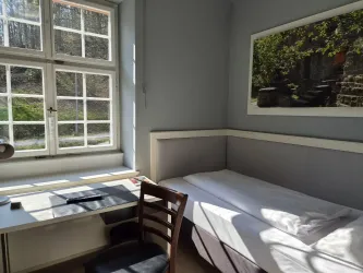 Ein schlichtes Zimmer mit einem Einzelbett und einem Schreibtisch. Der große Fenster lässt viel Tageslicht herein und bietet einen Blick ins Freie.