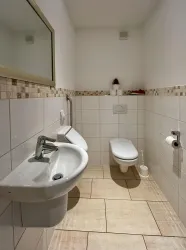 Ein modernes Badezimmer mit weißen Fliesen und einem Waschbecken. Rechts befindet sich eine Toilette und ein Spiegel über dem Waschbecken.