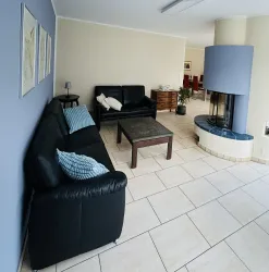 Ein gemütlicher Wohnraum mit schwarzen Sofas und einem runden Kamin. Der Raum hat helle Fliesen und eine entspannte Atmosphäre.