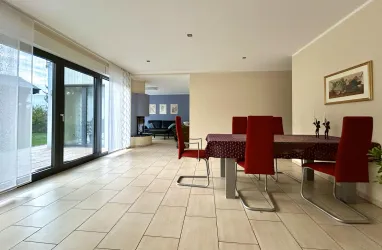 Ein heller Essbereich mit einem Tisch und roten Stühlen. Große Fenster lassen viel Licht herein und bieten einen Blick ins Freie.