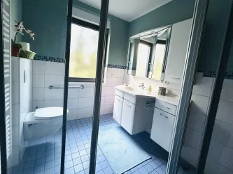Ein modernes Badezimmer mit einem weißen WC und einem hellen Waschbeckenmöbel. Die Wände sind in sanften Farbtönen gehalten und große Fenster sorgen für viel Tageslicht.