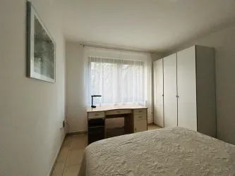 Ein einfaches Schlafzimmer mit einem Schreibtisch und einem großen weißen Kleiderschrank. Das Fenster lässt viel Licht herein und hat weiße Vorhänge.