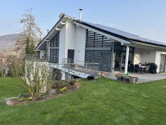 Ein modernes Haus mit einem großen Garten und einer Terrasse. Im Hintergrund sind sanfte Hügel und ein klarer Himmel zu sehen.