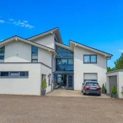 Ein modernes Haus mit großen Fenstern und einer eleganten Fassade. Vor dem Eingang parkt ein Auto in einer gepflasterten Auffahrt.
